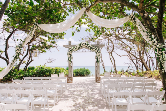 An outdoor beach wedding setup with white chairs, an aisle covered in flower petals, and an archway decorated with white flowers and draped fabric, surrounded by trees with a view of the ocean in the background.