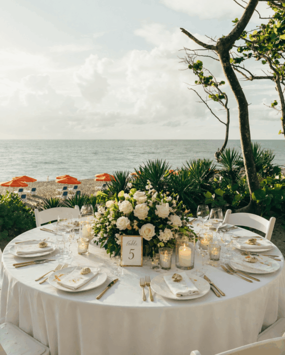 A round table set for a formal outdoor meal is decorated with white flowers, greenery, candles, and a table number 5 card, overlooking a beach with orange umbrellas and the ocean in the background.