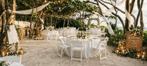 Outdoor beach wedding reception under trees with white drapery, string lights, round tables with white linens and chairs, floral centerpieces, and candles. A wooden welcome sign stands at the entrance.