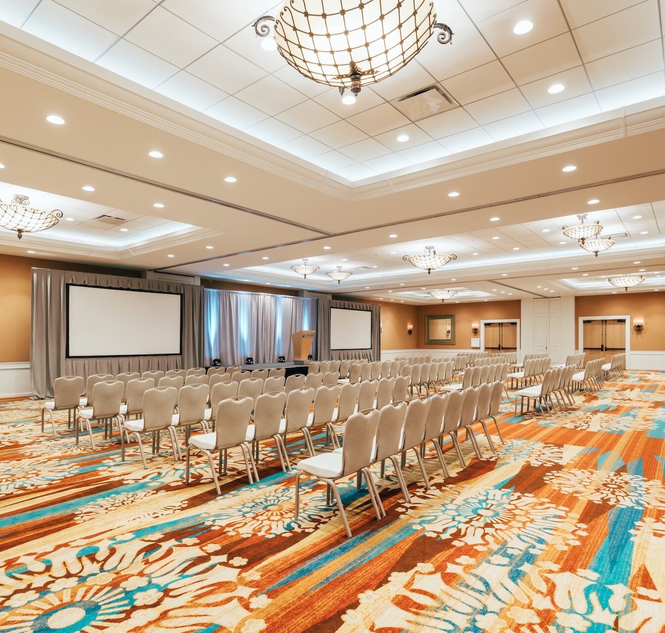 A spacious, brightly lit conference room with rows of beige chairs facing two large projection screens and a stage, decorated with colorful patterned carpet and elegant chandeliers.