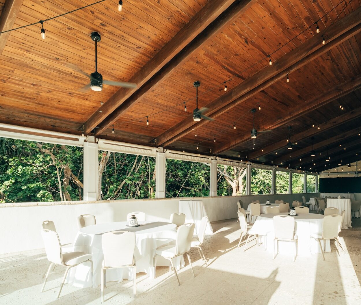 An outdoor covered event space with a wooden ceiling, string lights, ceiling fans, and round tables covered in white tablecloths, surrounded by white chairs. Sunlight streams in, and trees are visible outside the open walls.