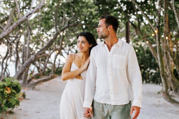 A couple walks hand in hand along a sandy path surrounded by trees and string lights. Both are dressed in light, summery clothing and appear happy and relaxed.