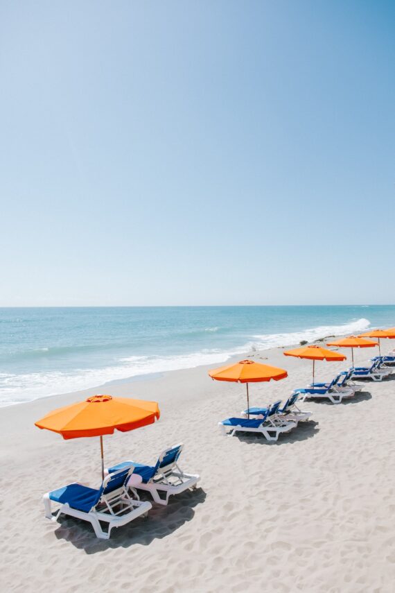 A row of blue lounge chairs with bright orange umbrellas is set up on a sandy beach facing the calm ocean under a clear blue sky.