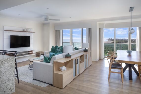 Bright, modern living and dining area with large windows overlooking the ocean. Room features a white sofa with teal pillows, wall-mounted TV, a wooden dining table, and light wood flooring.
