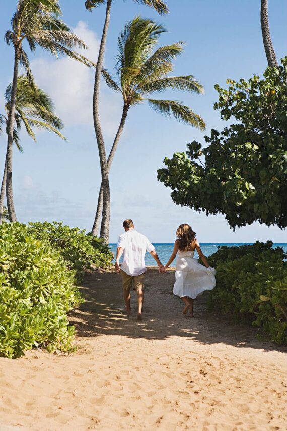 A couple holding hands walks along a sandy path toward the beach, surrounded by green bushes and tall palm trees, with blue sky and ocean in the background.