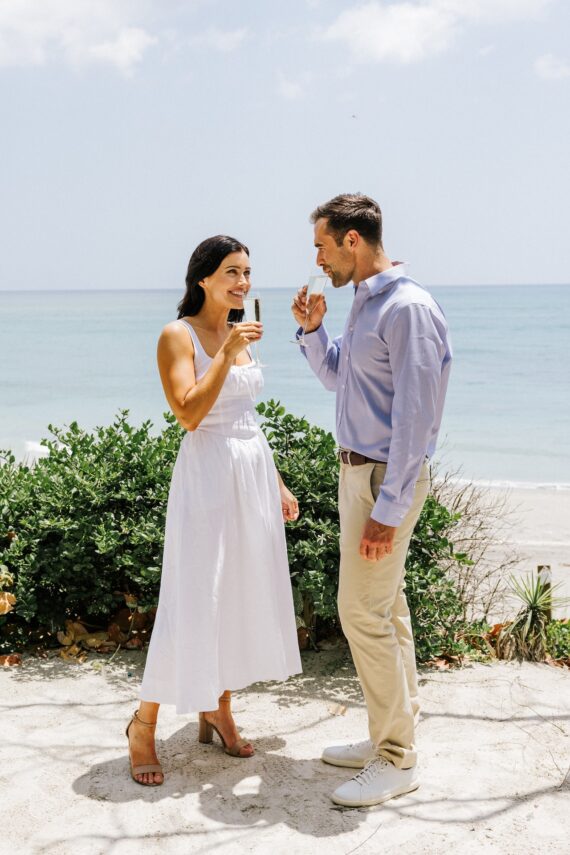 A woman in a white dress and a man in a light blue shirt and beige pants stand on a sandy beach, smiling and holding drinks, with the ocean and greenery in the background.