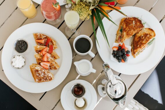 A table set for brunch with waffles topped with strawberries and powdered sugar, grilled sandwiches with fresh berries, coffee, juice, a smoothie, whipped cream, syrup, and butter on white plates.