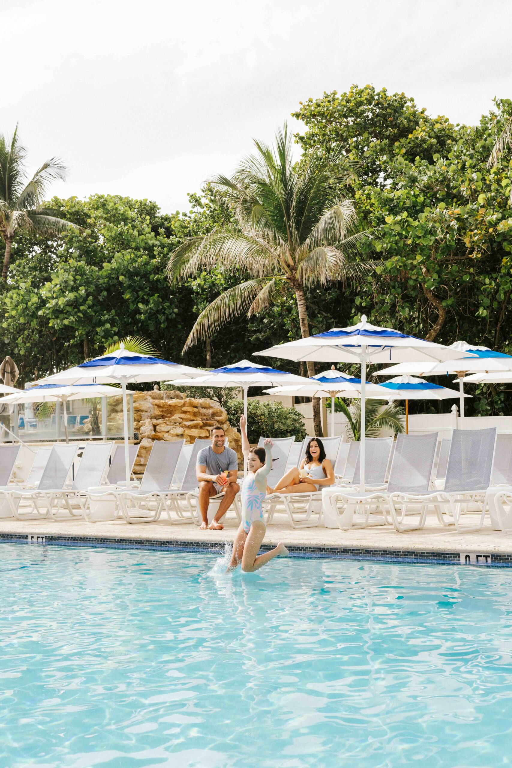 A child jumps into a swimming pool while two adults sit on lounge chairs nearby, surrounded by palm trees and white umbrellas.